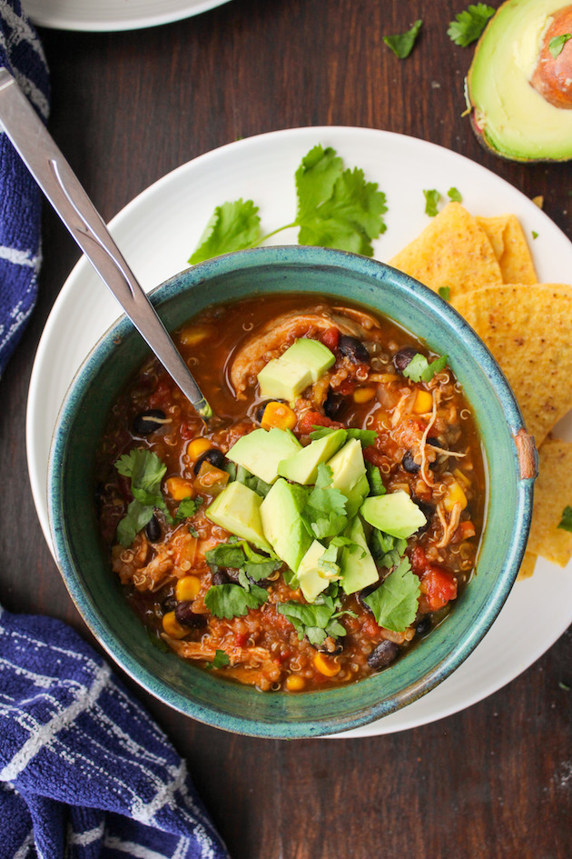 Instant Pot soup recipe for Spicy chicken enchilada soup - bowl of hearty chicken enchilada soup loaded with quinoa, beans, corn and garnished with chopped parsley and avocado, on a big white plate with corn chips on the side.