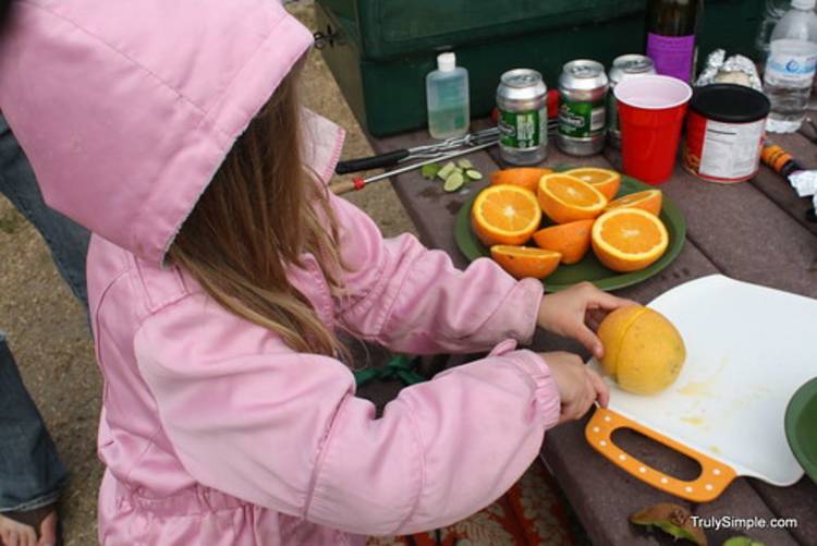 cold-weather-camping image of a child in a jacket cutting an orange on a cutting board at a campsite picnic table