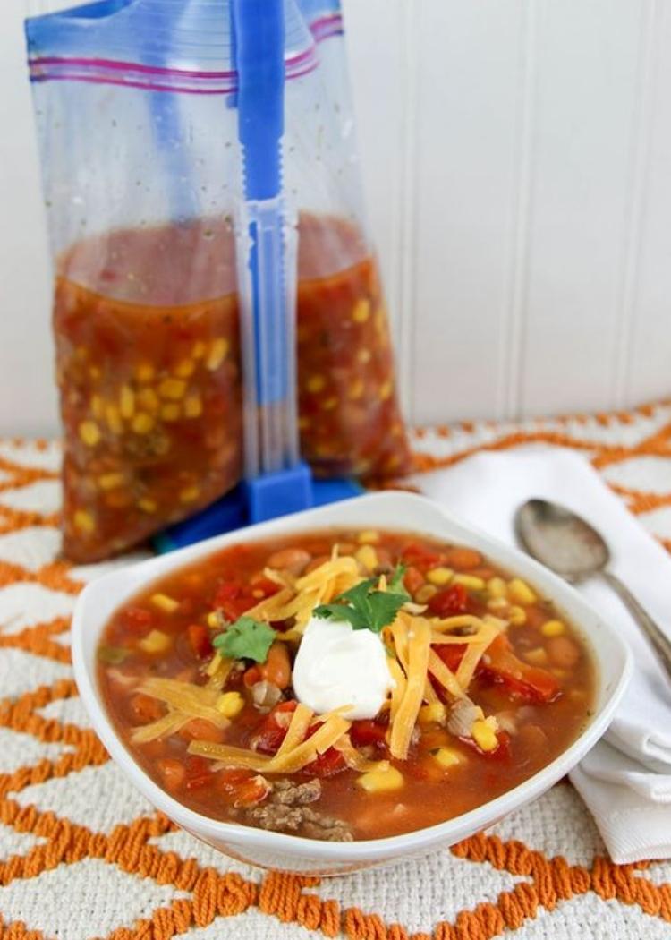 Taco soup - small bowl of taco soup with lots of cheese, sour cream and parsley garnish on top. With a freezer bag of taco soup in the background.