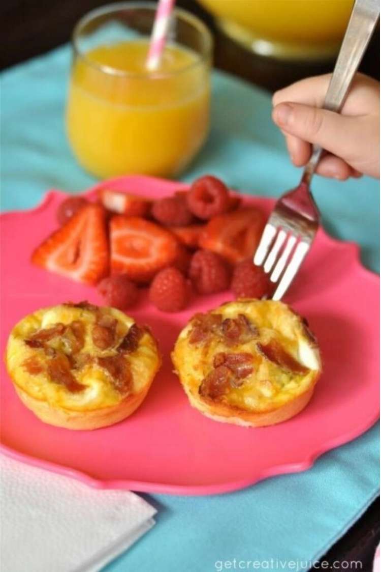 Plate with bacon and egg muffins and strawberries, hand with fork about to take bite, glass of orange juice in background