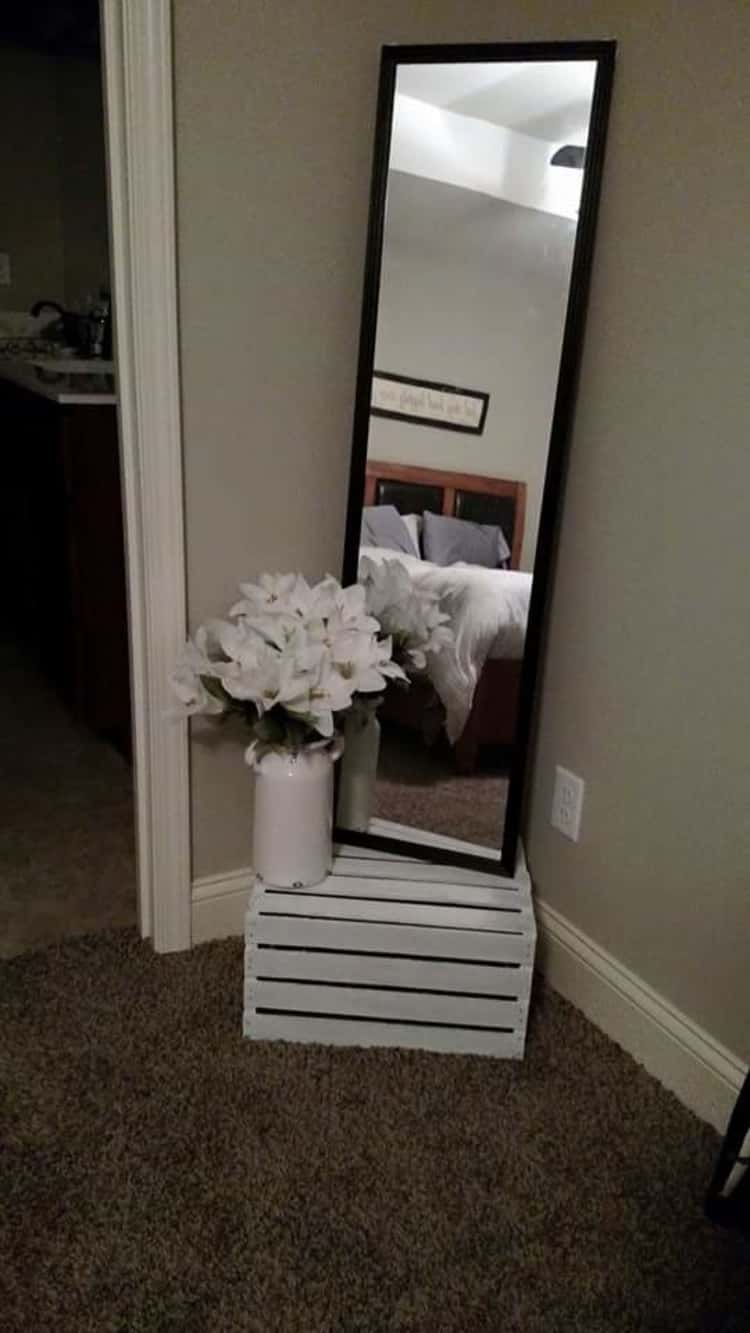 full length mirror in the corner sitting on a white crate with a vase of white flowers in front of it