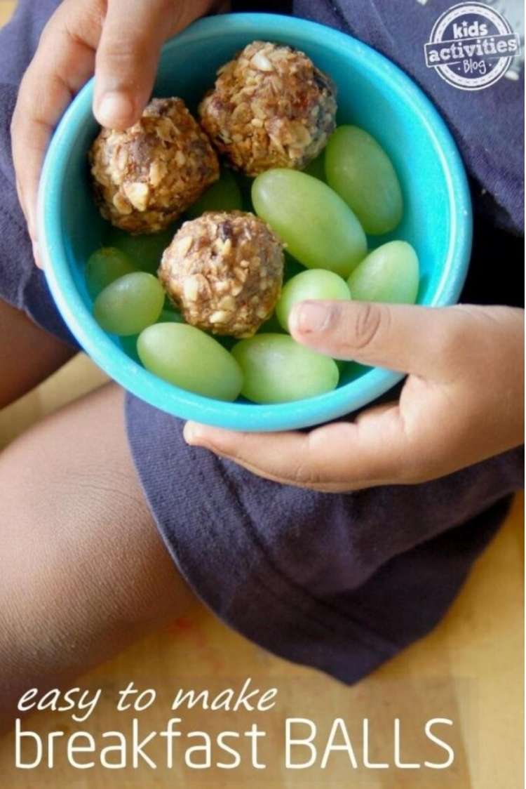 Child's hands holding a blue bowl containing several breakfast balls and fresh grapes
