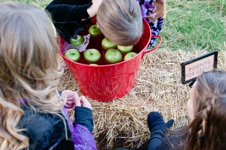 one kid bobbing her head for apples in a bucket filled with water and some apples while 2 other kids are watching